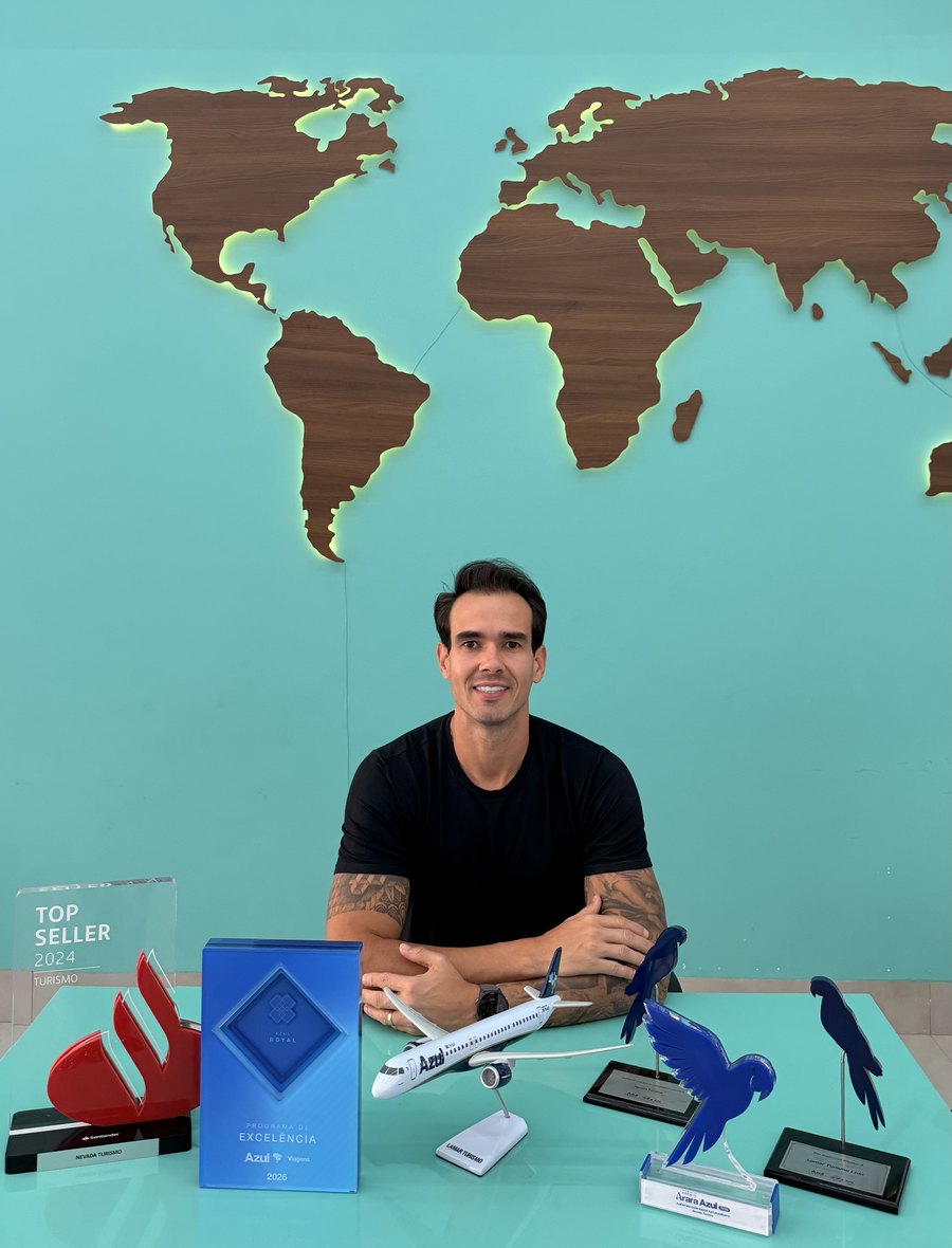 Bruno Augusto na sede da Nevada Turismo, diante do mapa-múndi em madeira, com os prêmios da agência sobre a mesa: Top Seller 2024 Turismo, troféu Santander, Programa de Excelência Azul Royal e os troféus Arara Azul